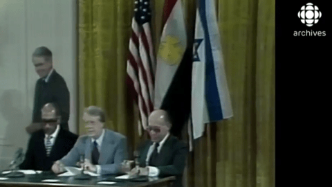 Les présidents Sadate et Carter, qu'accompagne le premier ministre Begin,  sont assis devant les drapeaux américain, égyptien et américain, pour signer les accords de Camp David.