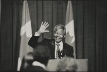 A black and white photograph of Nelson Mandela standing in front of a podium. He is smiling and waving.