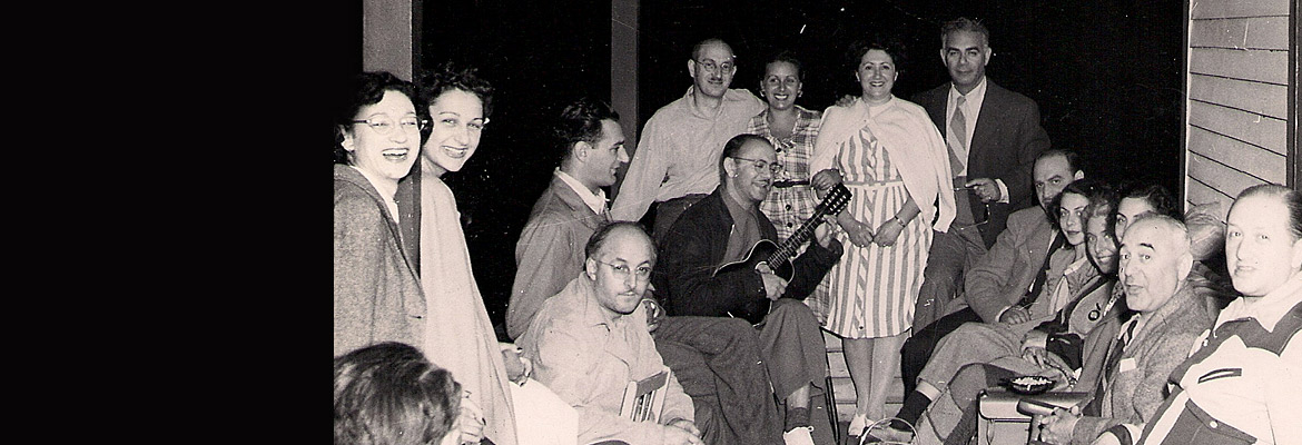 Un groupe de hommes et femmes assis sur une véranda en bois. Un des hommes tient un ukulélé.