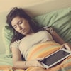Une jeune femme dort un lit avec sa tablette électronique entre les mains.