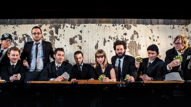 9 personnes habillées en noir, 8 hommes et 1 femme, posés devant la caméra derrière une table, certains avec des cartes à jouer dans les mains.