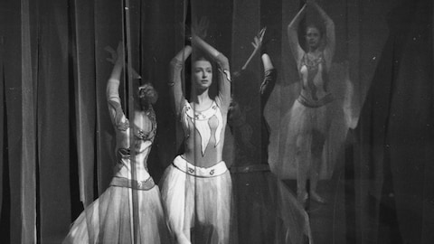Trois ballerines derrière des rideaux vaporeux dansent les bras levés. 