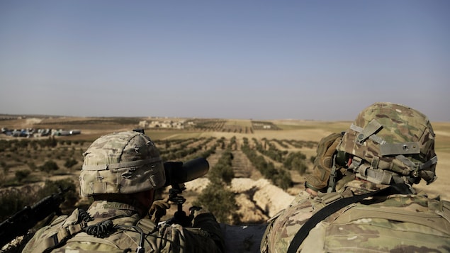 Deux soldats américains installés en hauteur regardent au loin, à l'aide de jumelles, le village de Manbij, au nord de la Syrie.