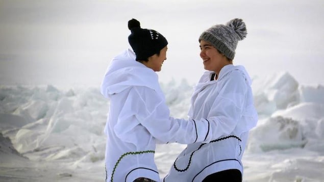 Les deux jeunes femmes sont face à face et se tiennent par les avant-bras, souriantes. Derrière elle, on voit un paysage enneigé et glacé.