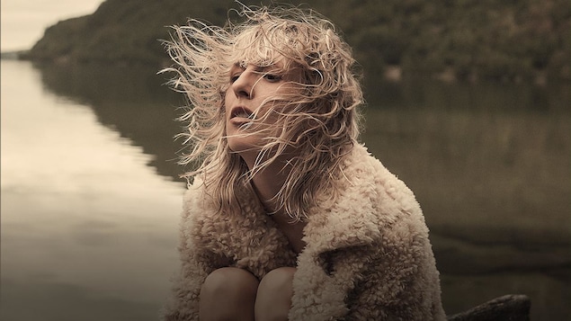 La chanteuse Ingrid St-Pierre assise aux abords d'un lac, on voit une montagne derrière; le ciel est gris. Ingrid porte un manteau qui ressemble à de la laine de mouton.