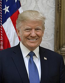 Head shot of a smiling Trump in front of an American flag. He is wearing a dark blue suit jacket, white shirt, light blue necktie, and American flag lapel pin.