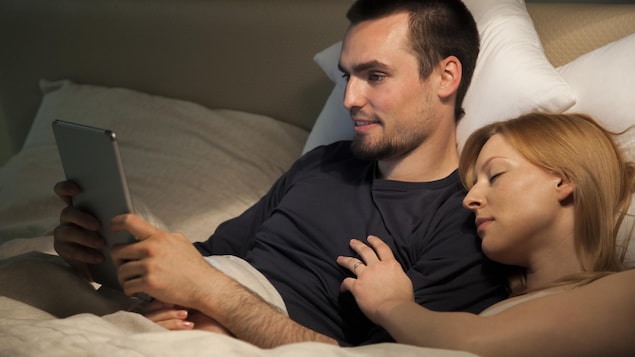 Un homme consulte sa tablette alors que sa femme dort à côté de lui.