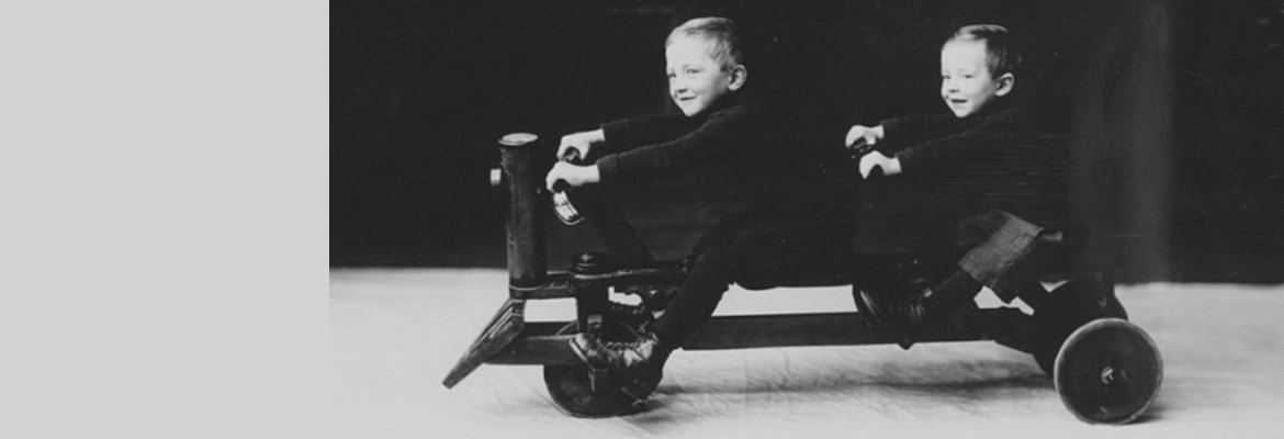 An old-fashioned black and white photograph of 2 smiling children seated on a cart that has 3 wheels and 2 sets of handlebars.