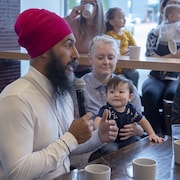 Jagmeet Singh parle dans un micro, assis à une table à côté d'une femme qui tient un bébé sur ses genoux.