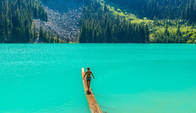Une femme en costume de bain marche en équilibre sur un billot de bois qui est dans un lac turquoise entouré d'une forêt.