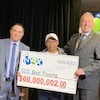Trois hommes tiennent un gros chèque sur lequel est écrit 60 000 002 $.