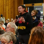 Un homme avec un enfant sur le dos parle dans un micro au milieu d'une salle remplie de gens.