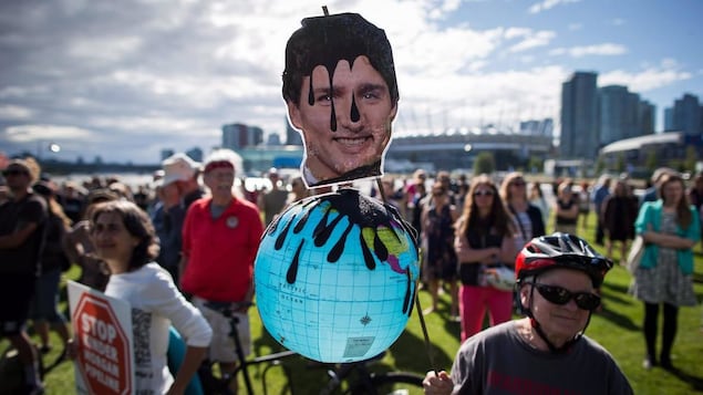 Un homme âgé tient un globe terrestre avec le visage de Justin Trudeau couvert en partie de pétrole noir. Des manifestants sont en arrière-plan devant un centre-ville. 