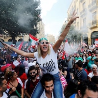 Une femme, sur les épaules d'un homme, tient un drapeau libanais. Tout autour d'elle, une foule de manifestants. 