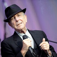 Le chanteur porte un chapeau noir et un complet, micro en main, sur une scène extérieure.