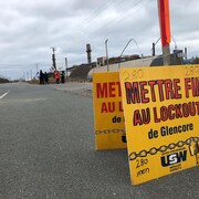 Une affiche placé sur la route devant la fonderie de Belledune. On peut y lire : « Mettre fin au lockout de Glencore ». Les grévistes marchent un peu plus loin. 