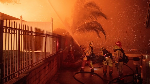 Des pompiers arrosent une maison tandis que des tisons volent autour d'eux.