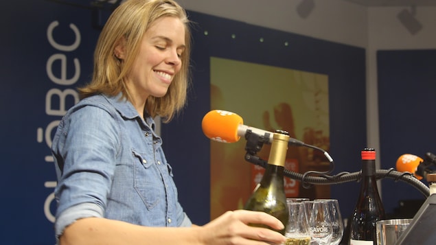 Une femme aux cheveux blonds et en chemise de jean tient une bouteille de vin à la main et remplit une coupe.