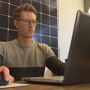 Dans un bureau, un homme l'air sérieux travaille avec un ordinateur portable avec le logo « Roots Rock Solar ». Derrière lui se trouve un panneau solaire.