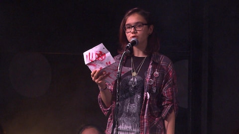 Une jeune adolescente portant des lunettes lit une lettre, debout, face à un micro.