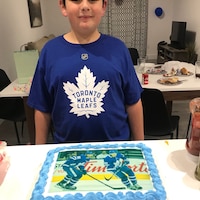 Un jeune garçon brun avec un t-shirt des Maple Leafs et un gâteau avec des joueurs de hockey en photo.