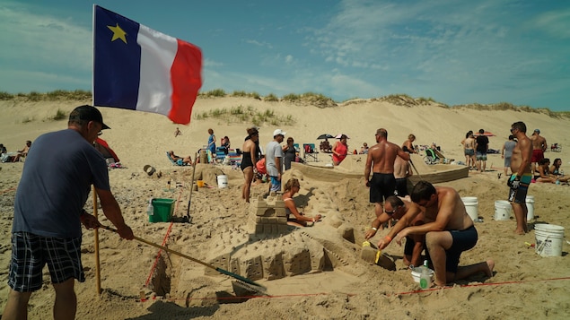 Un homme ratisse le tour de sa sculpture. Un drapeau acadien est planté dans le sable à côté.