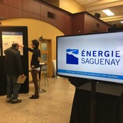 Une affiche d'Énergie Saguenay avec deux personne en arrière plan qui consultent des affiches. 