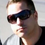 Un homme, proche de la quarantaine, avec un t-shirt noir et des lunettes noires. 