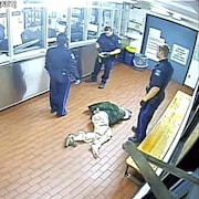 Un homme couché sur le sol menotté dans un poste de police, avec trois policiers debout autour de lui.