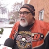 Un homme vêtu d'une tuque et d'un chandail Harley-Davidson parle au micro d'un journaliste à l'extérieur d'un immeuble.