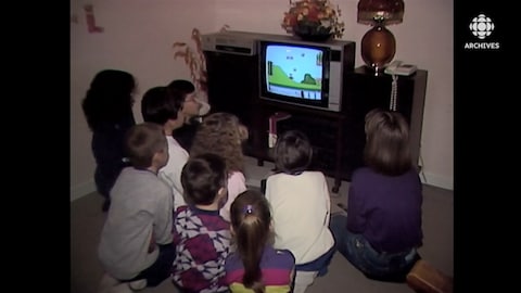 Un groupe d'enfants sont tournés vers un écran de téléviseur sur lequel se déroule une partie du jeu Mario Bros sur console Nintendo.