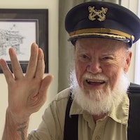 Dick Stevenson portant une casquette de capitaine de bateau sourit en saluant de la main.