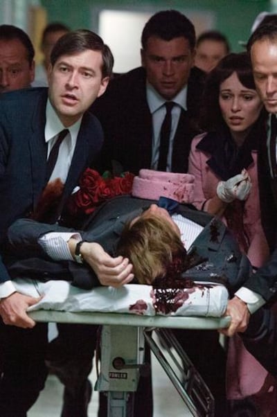 Un groupe d'hommes et une femme en tailleur rose autour d'une civière portant un corps ensanglanté dans un couloir d'hôpital.
