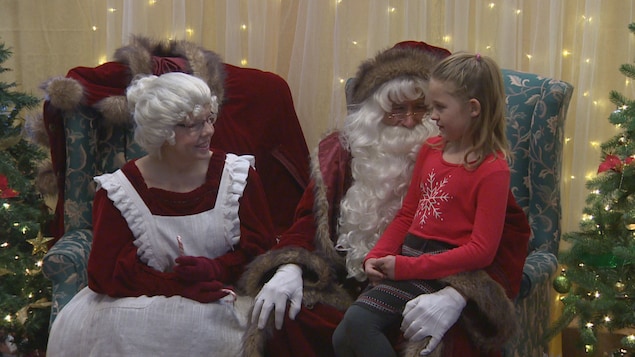Deux personnes déguisées en père et mère Noël. Une petite fille est assise sur les jambes de père Noël.