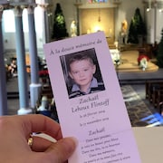 Une main tient un pamphlet en l'honneur de Zackaël lors de ses funérailles. 