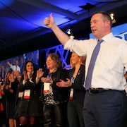 Jason Kenney salue ses militants d'un pouce levé, sous les applaudissements de ses ministres, debout derrière lui.