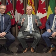 Les trois premiers ministres sont assis sur des chaises l'un à côté de l'autre devant des drapeaux canadiens et de leurs provinces respectives.