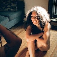 Une jeune femme avec les cheveux bouclés est assise sur un plancher de bois, sans vêtement. Elle sourit à la photographe devant elle, et le soleil plombe sur ses cheveux.