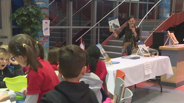 Danielle Loranger est debout avec son livre dans les mains. Elle présente son livre devant des enfants assis à une table.