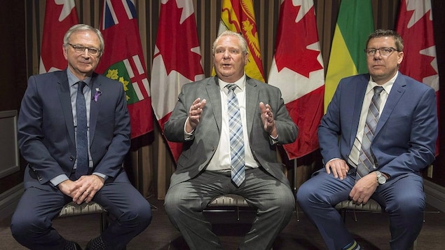 Les trois premiers ministres sont assis sur des chaises l'un à côté de l'autre devant des drapeaux canadiens et de leurs provinces respectives.