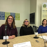 Trois femmes assises derrière une table avec des micros donnent une conférence de presse sur la violence conjugale en milieu de travail. 