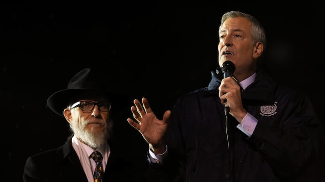 Bill de Blasio parle dans un micro. Un rabbin se trouve à ses côtés.