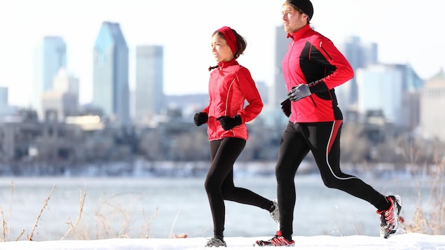 Une femme et un homme courent courent sur un sentier de neige. Les gratte-ciel de Montréal se trouvent en arrière-plan.