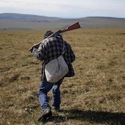 Plan large d'un chasseur vu de dos marchant avec une arme sur l'épaule dans une grande plaine.