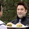 Le comédien sourit en tenant un plateau avec deux sandwiches. 