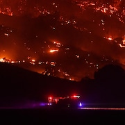 Un camion des pompiers passe devant un flanc de montagne en proie aux flammes dans la nuit.