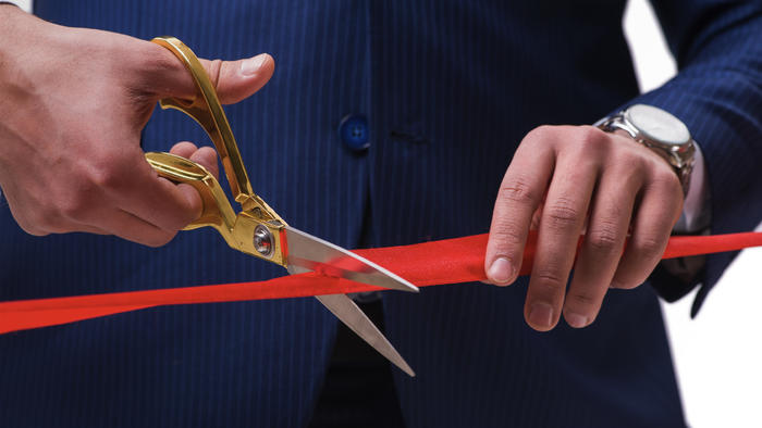 A close up of a man wearing a suit cutting red tape with scissors.