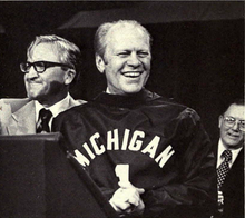A black and white image of an older man wearing a sweatshirt with the word "Michigan" on it standing on a stage.