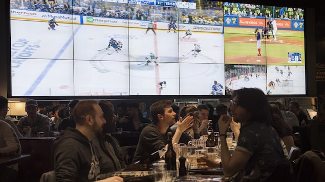 Des partisans discutent autour d'une table dans un bar pendant que des événements sportifs sont diffusés sur un écran géant.