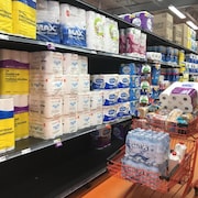 Des étagères avec du papier de toilette et un panier d'épicerie rempli de bouteilles d'eau, de papier de toilette et de pain.